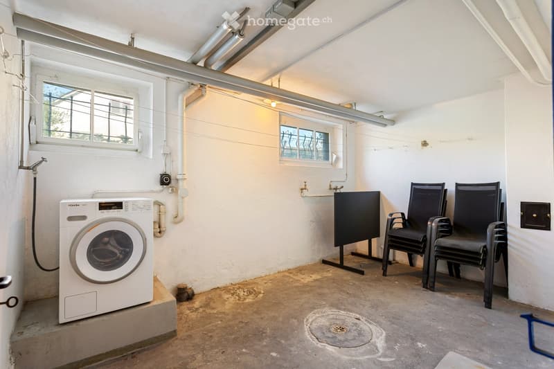 Utility room with a washing machine, two black chairs, and a screen, featuring exposed pipes and windows.