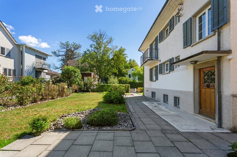 A well-maintained garden area with a paved pathway leading to a house entrance, surrounded by greenery and shrubs.