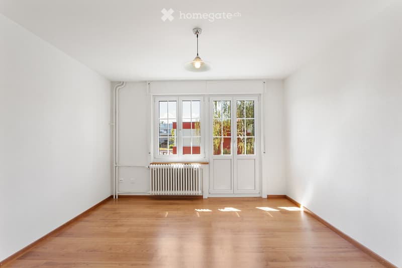 Empty room with white walls, wooden floor, large windows, radiator, and a single hanging light fixture.