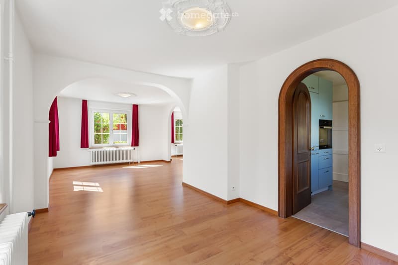 Empty room with wooden flooring, white walls, large windows with red curtains, and a radiator. An arched doorway leads to a kitchen area with blue cabinets and stainless steel appliances.