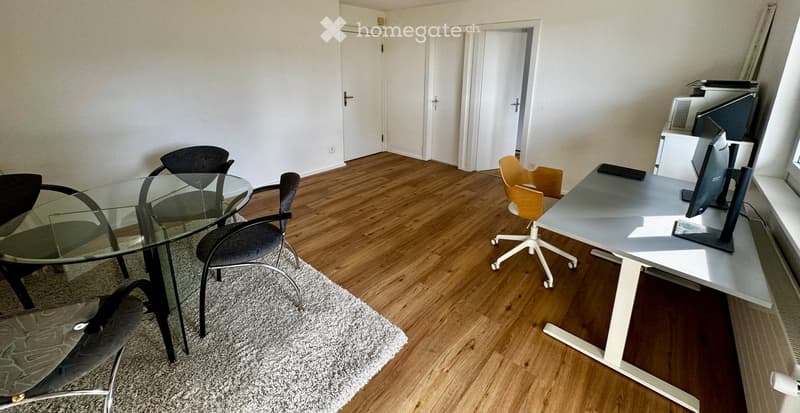 Modern office space with wooden flooring, glass table, office chair, desk with computer monitor, and additional chairs.
