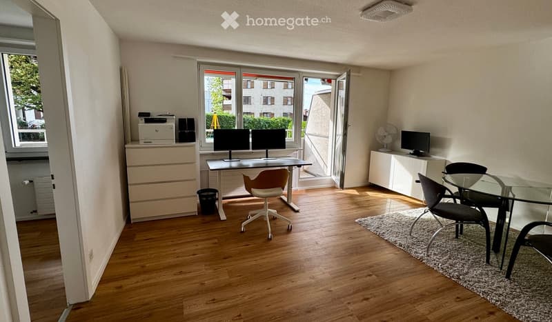 Bright, modern room with wooden flooring, large windows, and a balcony. Contains a desk with two monitors, a chair, a small table with chairs, a printer, and a fan.