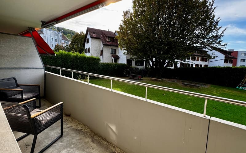 Balcony with two chairs and a small table, overlooking a green lawn and residential houses.
