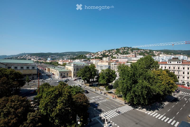 A panoramic view of a cityscape with buildings, roads, and greenery, featuring a clear blue sky and distant hills.