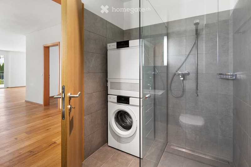 Modern bathroom with a glass-enclosed shower, a toilet, and a stacked washing machine and dryer unit.