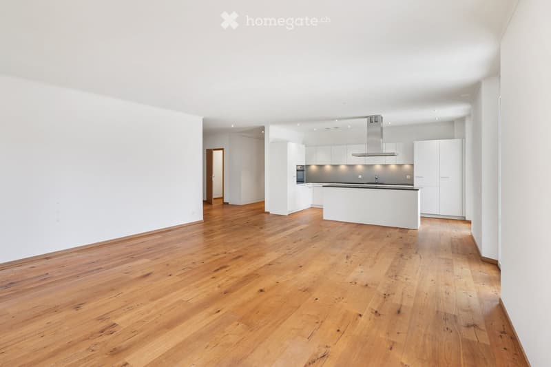 Open-plan unfurnished room with light wooden flooring, white walls, and a modern kitchen area featuring white cabinets, a stainless steel range hood, and a black countertop.
