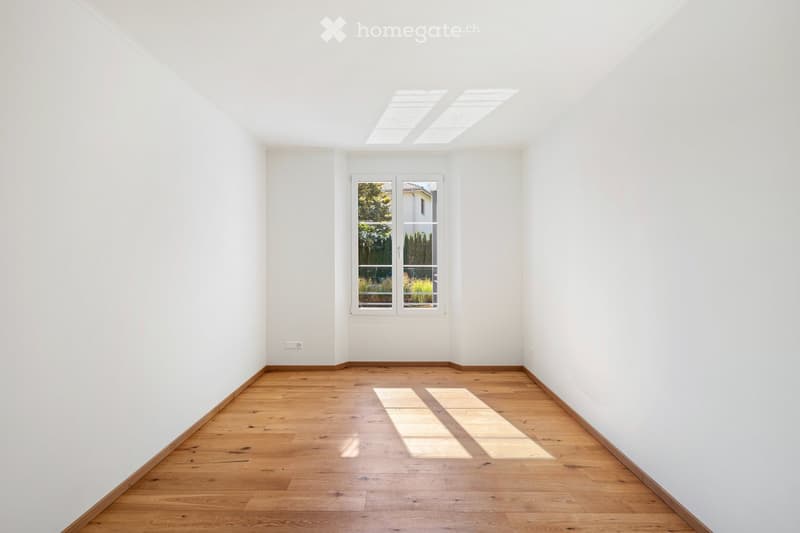 Empty room with white walls, wooden floor, large window, and skylights allowing natural light to enter.