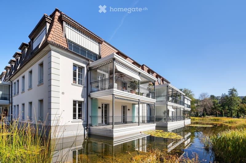 A modern, multi-story house with white walls, large windows, and a tiled roof, situated on stilts above a reflective water body with lily pads and surrounding greenery.
