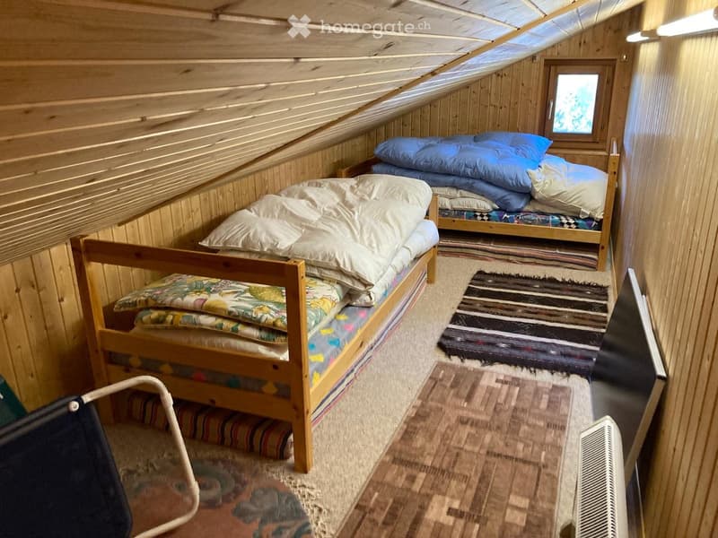 Cozy attic bedroom with wooden walls and slanted ceilings, featuring two wooden beds with colorful bedding, a small table, a chair, and a screen.