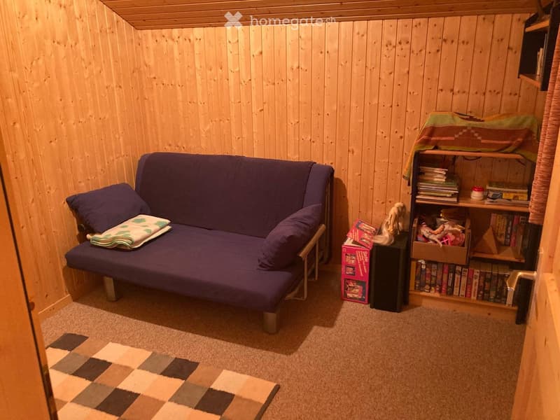 A small, cozy room with wooden walls and a purple futon sofa, a bookshelf filled with books, a small table with a toy box, and a blonde doll on top.