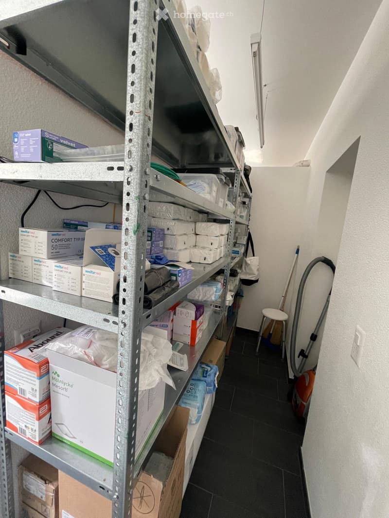 A storage room with metal shelving units filled with various boxes and items, a small white stool, and a vacuum cleaner leaning against the wall.