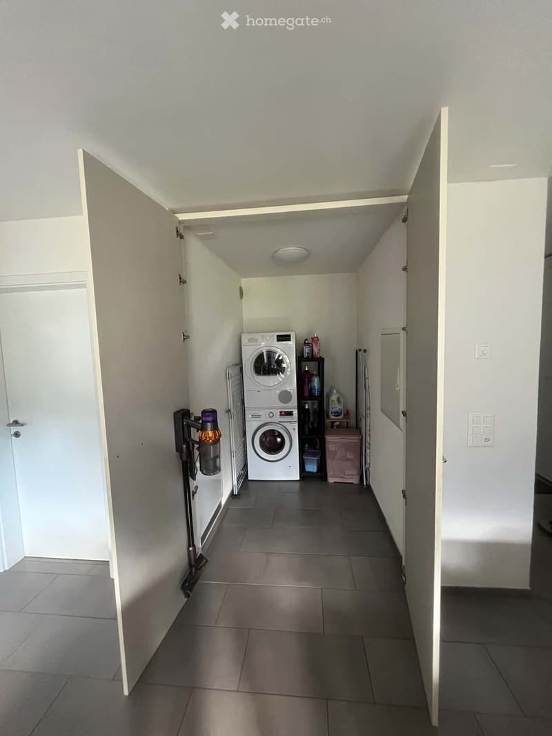 A narrow hallway with tiled flooring and white walls, leading to a laundry area with a washing machine, dryer, and cleaning supplies.