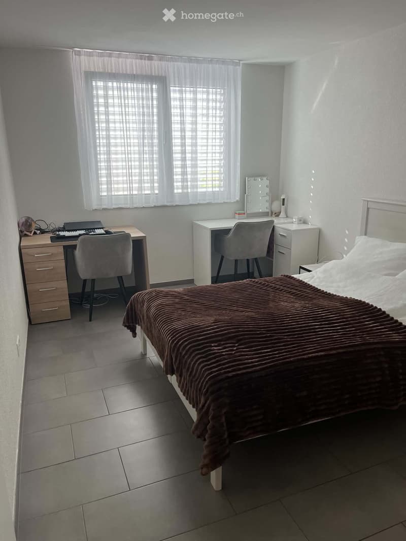A modern bedroom with a large bed, a desk with a laptop and keyboard, a vanity with a mirror, and two chairs.