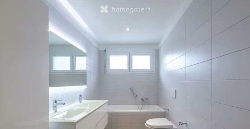 Modern, minimalist bathroom with white tiles, two sinks, a bathtub, and a toilet. Features large windows and sleek lighting.
