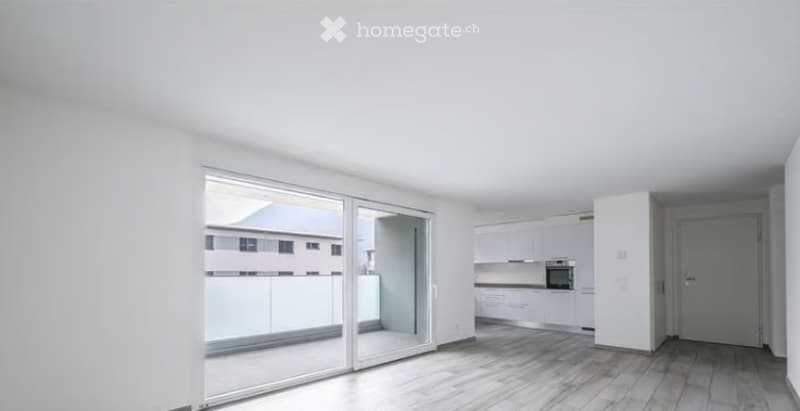 Bright, empty room with large sliding glass doors leading to a balcony, white walls, light wooden flooring, and a small adjacent kitchen area with white cabinets and appliances.