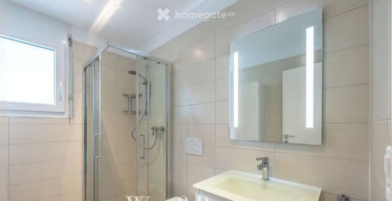 Modern bathroom with a white tiled shower, a large illuminated mirror, and a white sink.