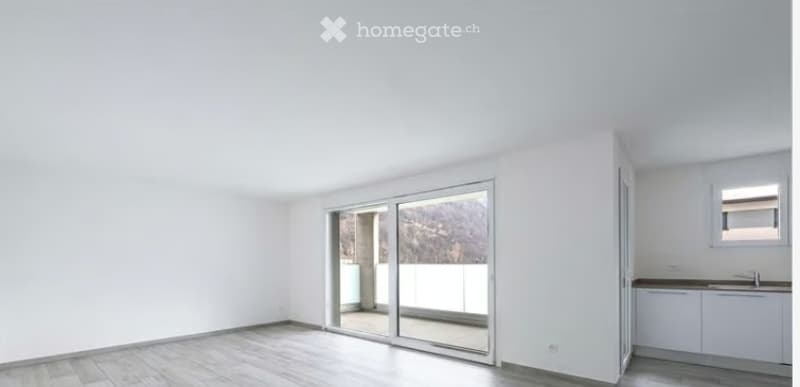 Empty room with white walls, wooden floor, large sliding glass doors leading to a balcony, and a small kitchenette area with white cabinets and a sink.