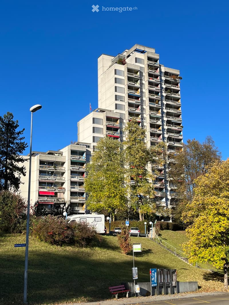 Multi-story residential building with balconies, surrounded by trees and a grassy slope with a bench and parked vehicles.
