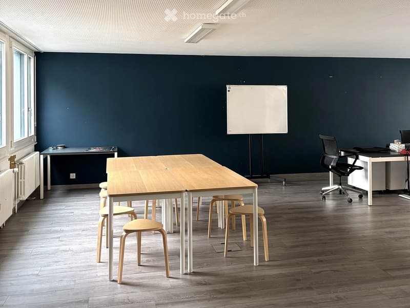 Modern office space with dark blue walls, light wooden flooring, and large windows. Features a long wooden table with multiple chairs, an office chair, a desk, and a whiteboard.
