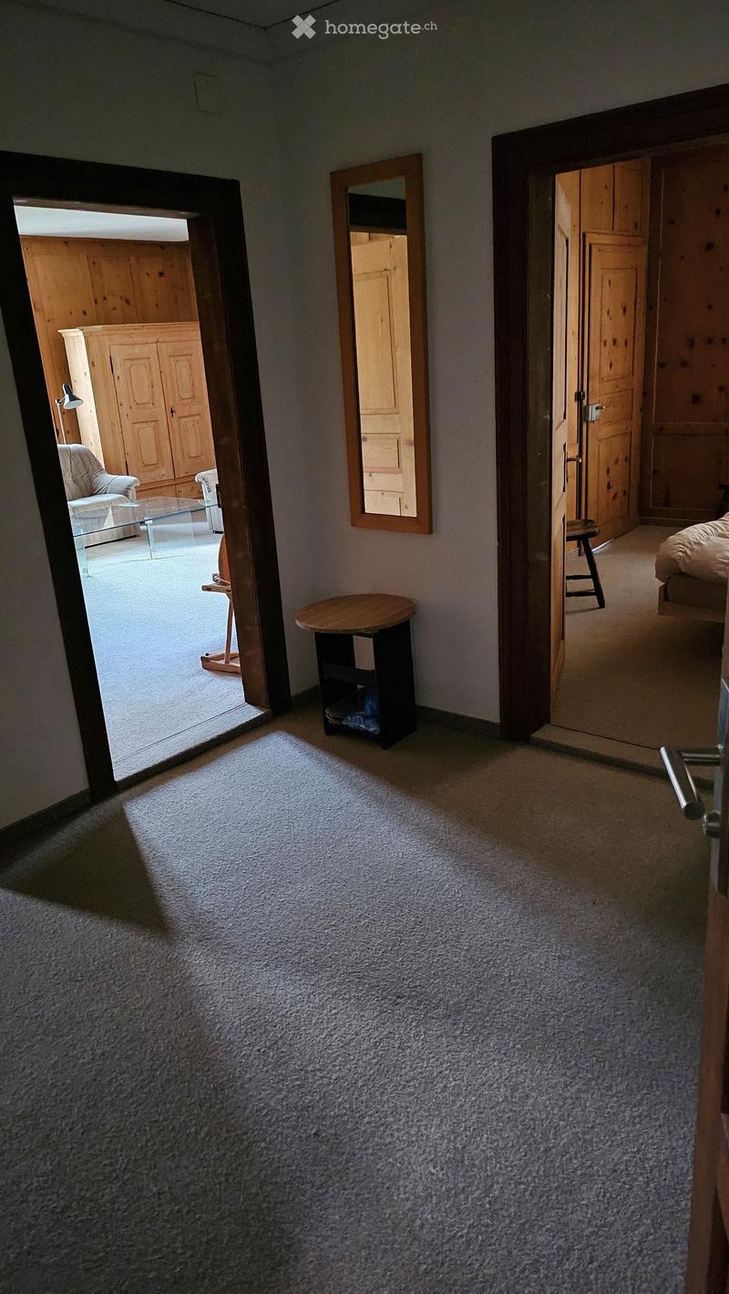 Carpeted hallway with open doors leading to a living room and bedroom, featuring a small round table and a mirror on the wall.