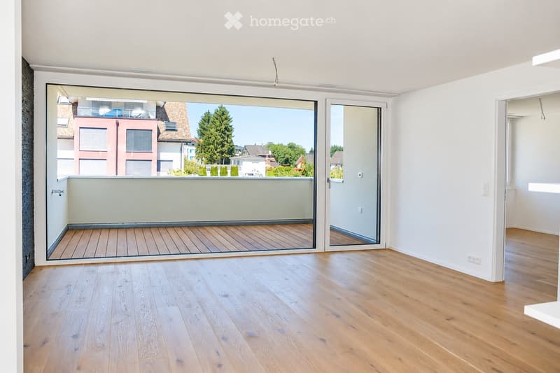 Empty room with light wooden flooring, large sliding glass doors leading to a wooden decked balcony, white walls, and modern lighting fixtures.