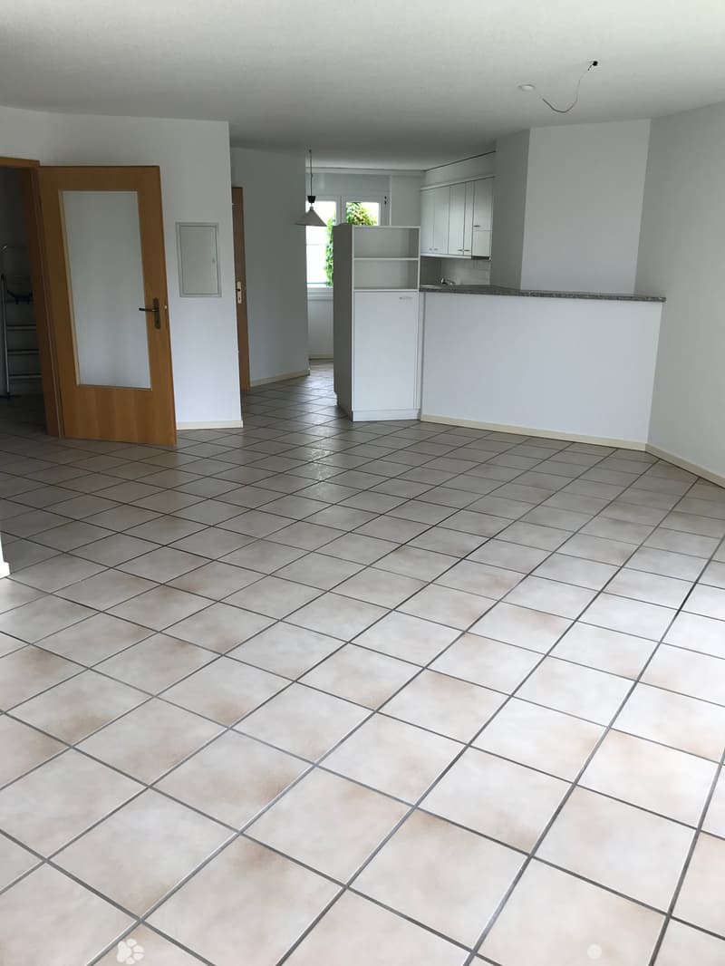 Empty room with tiled floor, white walls, and a doorway leading to another room. The adjacent area shows a small kitchen with white cabinets and a countertop.