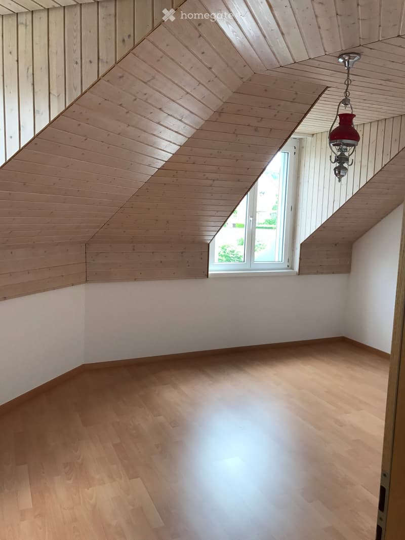 Empty room with wooden floor and slanted wooden ceiling, featuring a single window and a hanging red lantern light fixture.