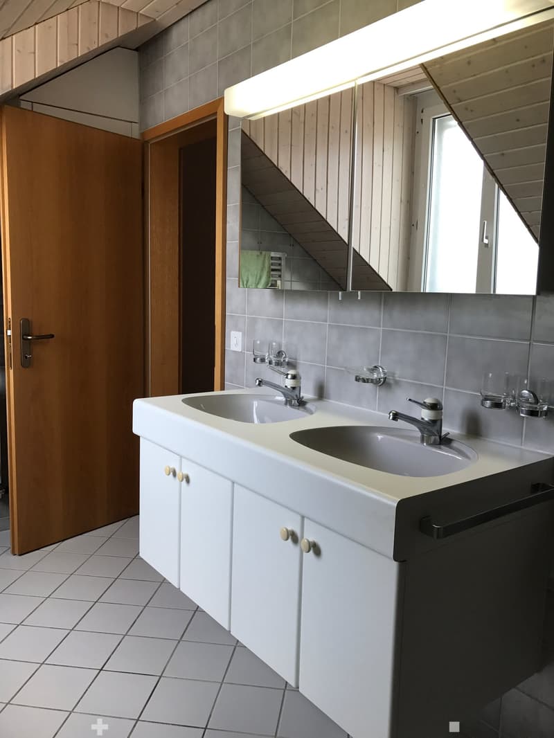 Modern bathroom with a double sink, white cabinets, and a large mirror above the sink. The room features tiled floors and walls, with a wooden door visible on the left side.