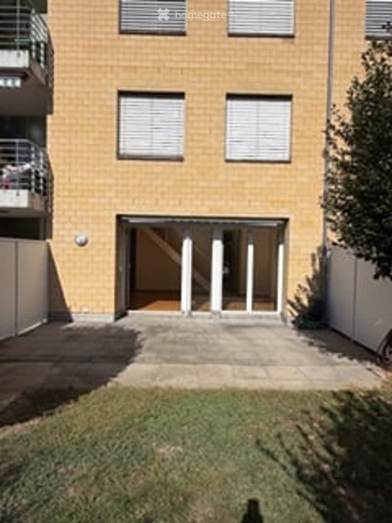 Modern multi-story building with a covered garage entrance, white columns, and a small grassy area in front.
