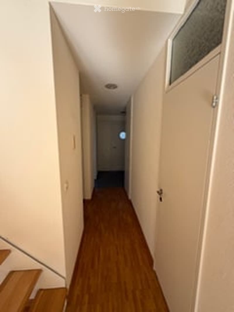 A narrow hallway with wooden flooring, white walls, and a small window at the end. The hallway leads to a door and has a staircase on the left side.