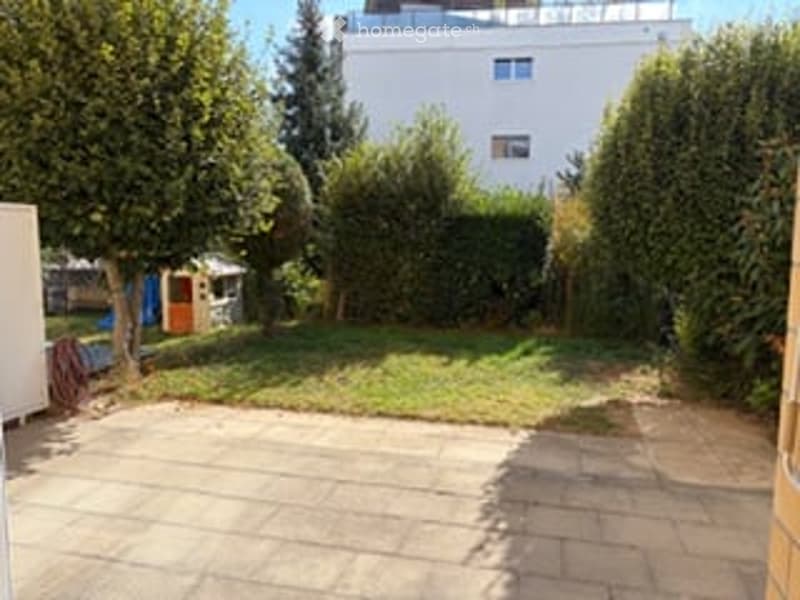 A well-maintained garden area with a paved patio, green lawn, and several trees and shrubs. A small playhouse is visible in the background.