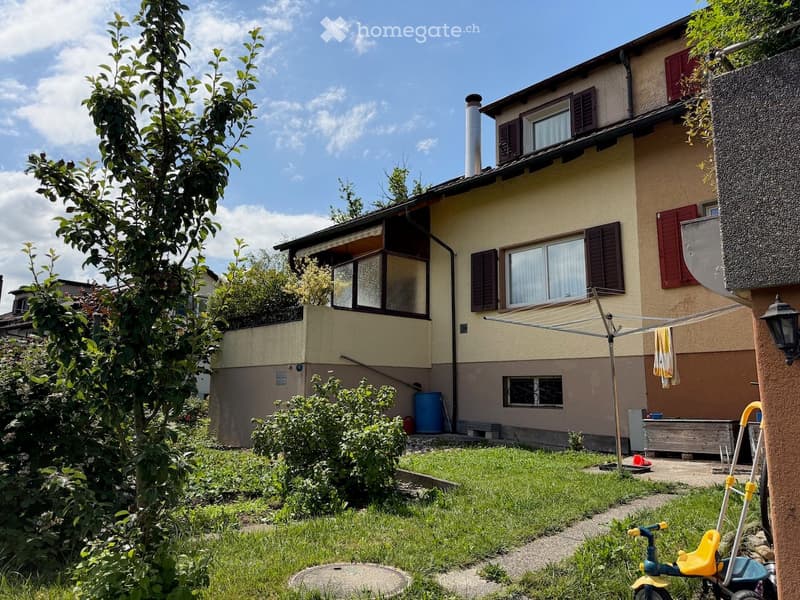 A well-maintained garden area in front of a two-story house with beige walls, red shutters, and multiple windows. The garden features a grassy lawn, small trees, shrubs, a clothes drying rack, a blue barrel, and a small tricycle.