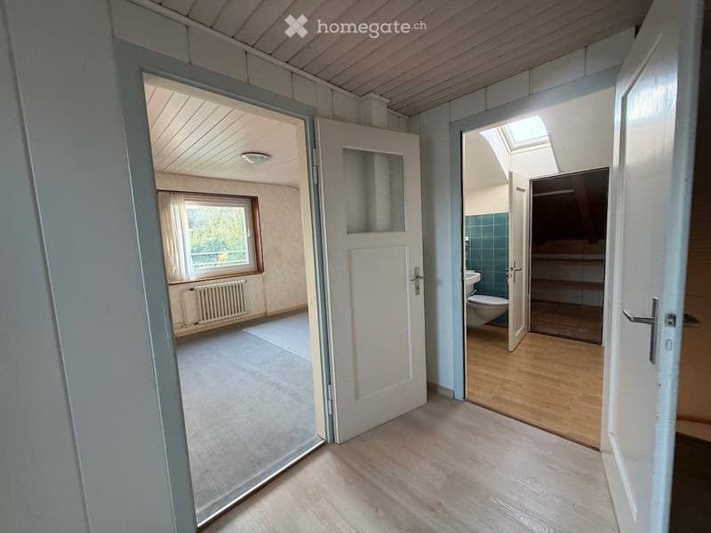 Empty hallway with wooden flooring, open doors leading to a room and a bathroom, visible window and radiator in the adjacent room.