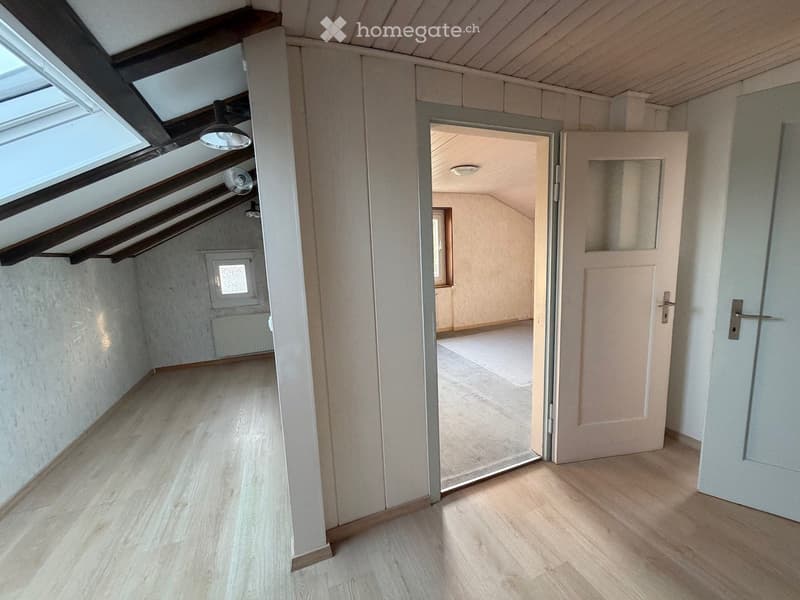 Empty hallway with wooden flooring, white walls, and exposed wooden beams on the ceiling. Features a skylight window and an open doorway leading to another room.