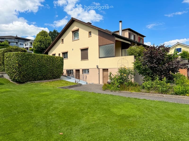 Two-story house with beige and brown exterior, multiple windows, a chimney, and a well-maintained lawn with surrounding greenery.