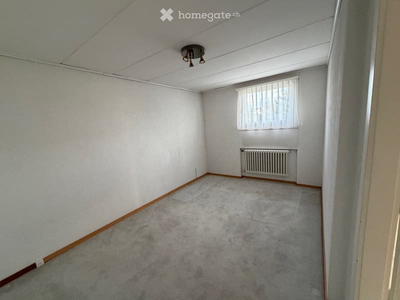 Empty room with white walls, grey carpet, a window with white blinds, a radiator, and a ceiling light fixture.