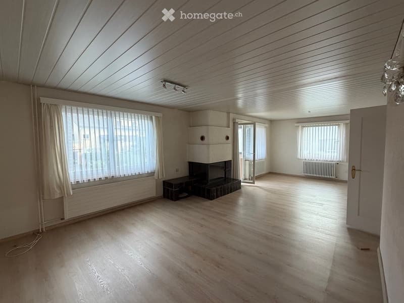 Empty room with wooden flooring, white walls, large windows with blinds, and a fireplace with a small cabinet.