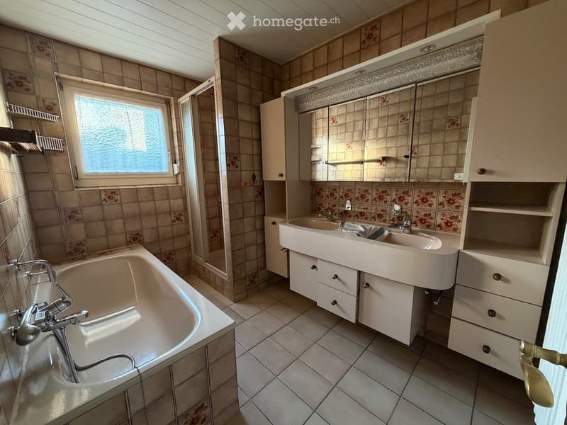 Bathroom with a bathtub, double sink, mirror, and cabinet. The walls are tiled with floral patterns, and there is a window with frosted glass.