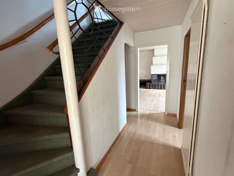 A well-lit hallway with wooden flooring, a staircase with a wooden handrail and metal balustrade, and an open doorway leading to another room.