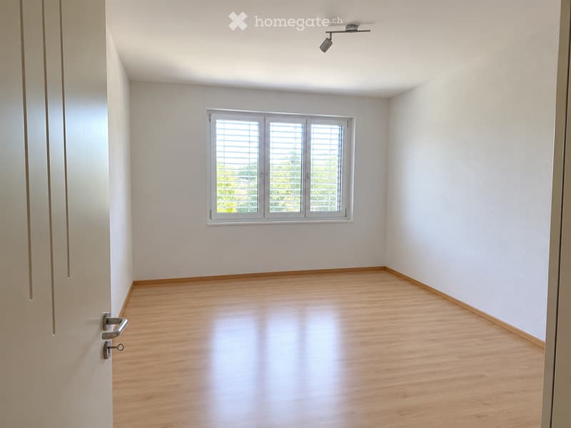 Empty room with light wooden flooring, white walls, and a large window with blinds. The room has a simple, modern design with a door visible on the left side.
