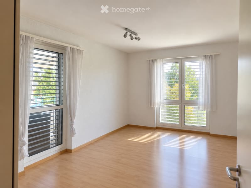 Empty room with light wooden flooring, white walls, large windows with white curtains, and a ceiling light fixture.