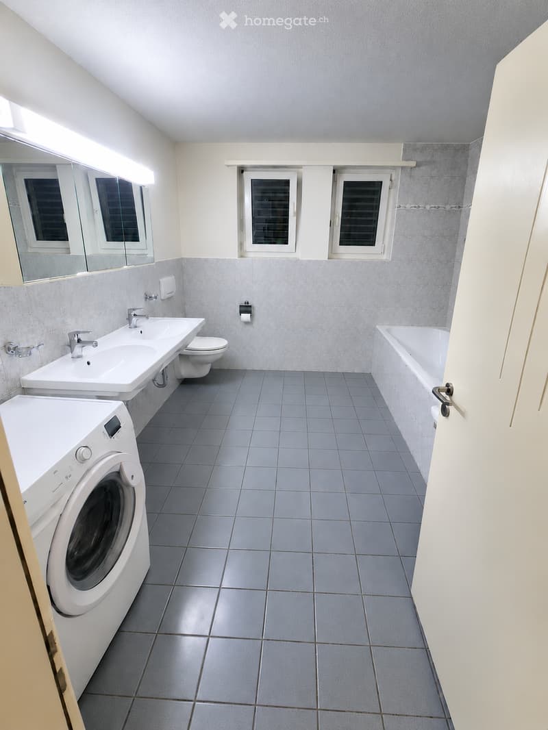 Modern bathroom with grey tiled floor, white sink, toilet, bathtub, washing machine, and large mirror.