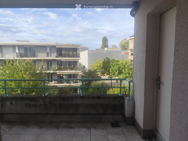 Balcony with tiled floor, green metal railing, overlooking a multi-story building and greenery.