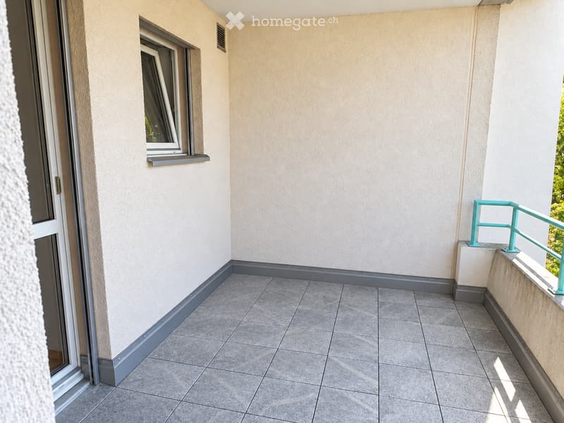 Balcony with tiled floor, light-colored walls, and a small window with a grey sill. Features a turquoise railing and a door leading inside.