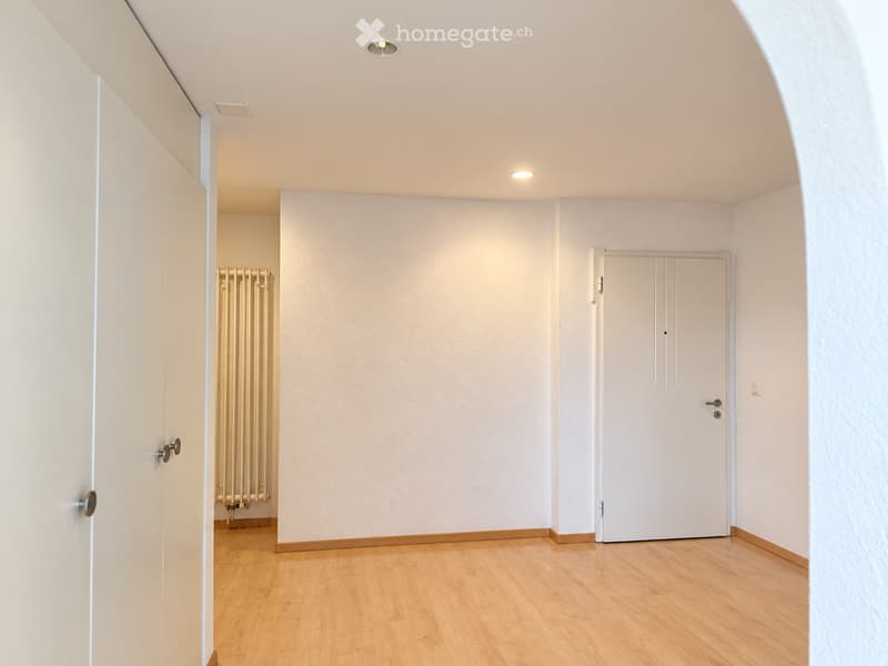 Empty room with light-colored walls, wooden flooring, and recessed ceiling lights. Features a radiator and two doors.