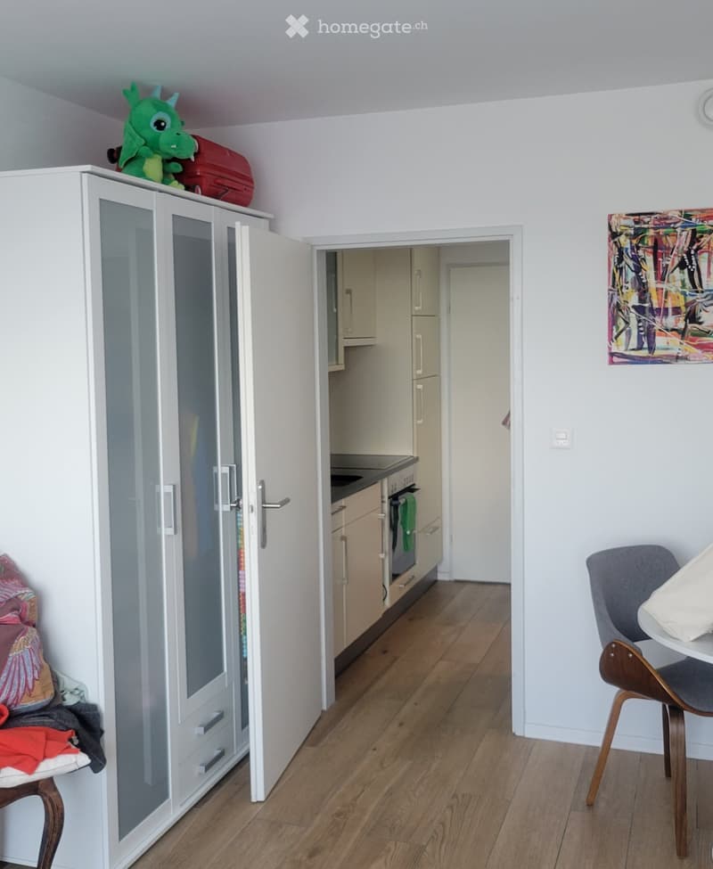 A furnished room with a wooden floor, a white wardrobe with glass doors, a small table with a chair, and a colorful painting on the wall. A small kitchen area is visible through an open doorway.