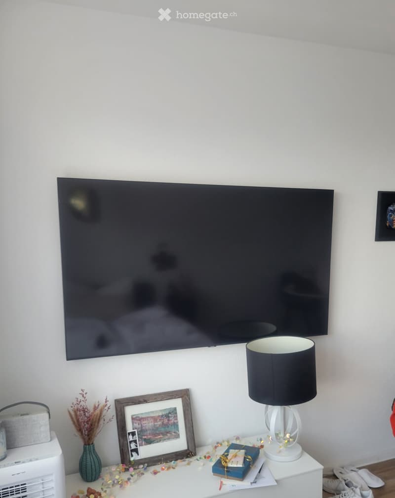 A modern living room with a large flat-screen TV mounted on the wall, a white side table with a lamp, a framed picture, a vase with dried flowers, and a small decorative arrangement.