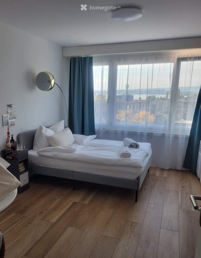 Cozy bedroom with a large bed, nightstand, floor lamp, and a scenic view through large windows.