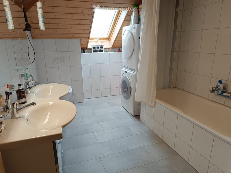 Bathroom with a double sink, bathtub, washing machine, and skylight window. The room features tiled walls and floor, and includes various toiletries and a mirror.