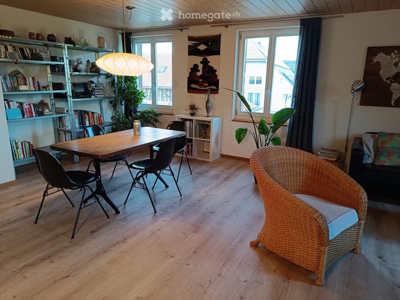 Bright living room with wooden flooring, large windows, a wicker chair, sofa, dining table with chairs, bookshelf, and decorative items.
